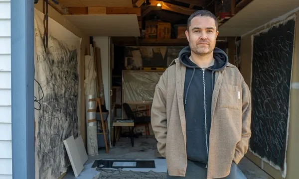 A man stands in front of a garage art studio, wearing a brown jacket and black hoodie. Behind him are large abstract artworks, canvases, and art supplies.