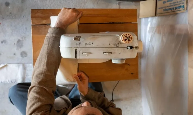 A person uses a white sewing machine on a wooden table, viewed from above. Fabric is being fed through the machine, and cardboard boxes are stacked nearby.