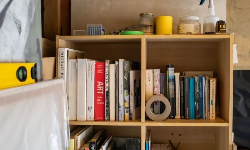 A wooden bookshelf filled with books, masking tape, a level, jars, and various small items. Some books are standing upright, while others are stacked. The background shows a partially painted wall.