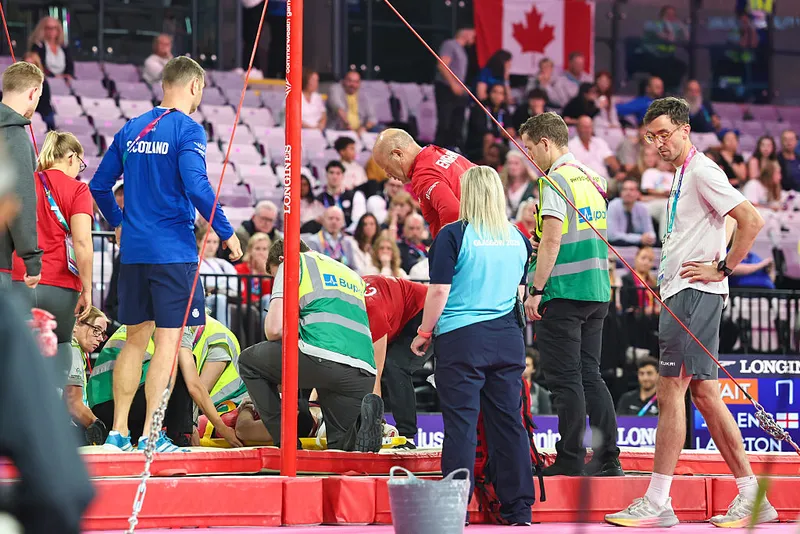 A group of medics and coaches gather around a young man lying flat on his back on a gymnastics mat as a crown looks on from the stands