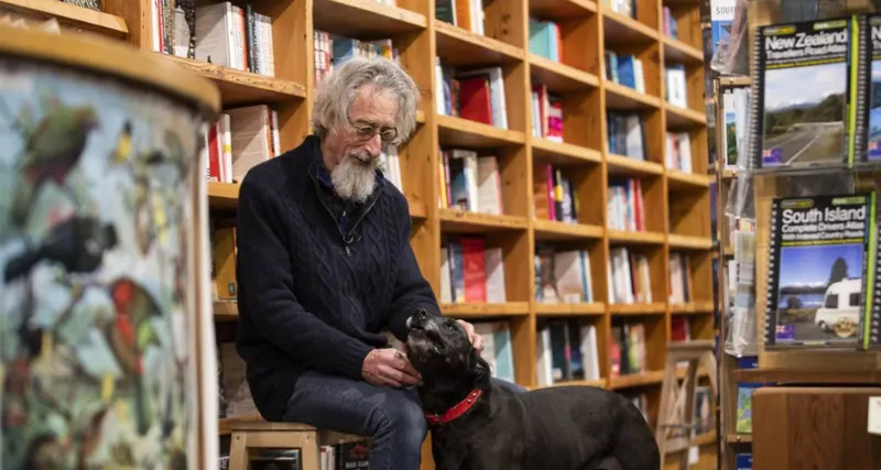 An older man with grey hair and beard, and wearing glasses is seated in a bookshop, patting a dog.