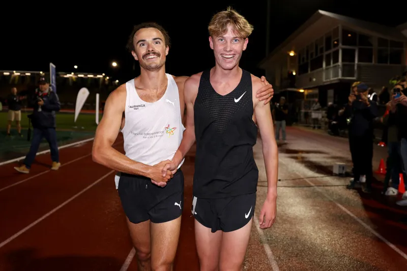 two lean young men wearing running shorts and singlets walk on a track shaking hands and smiling