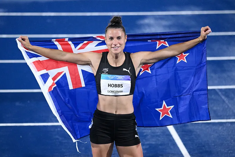 a woman wearing a balck running crop top and black running tights smiles on a blue track and holds a new zealand flag