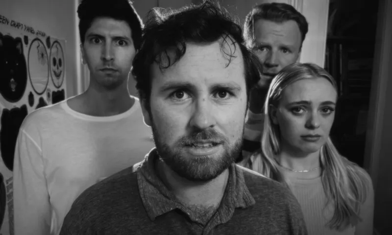 Four adults stand together indoors, facing the camera with serious or concerned expressions. The image is in black and white, and the background includes a wall with posters and a shelf.