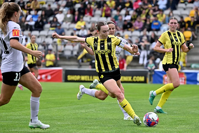 soccer players wearing white uniforms chase a female player in a striped black and yellow uniform as she kicks a ball