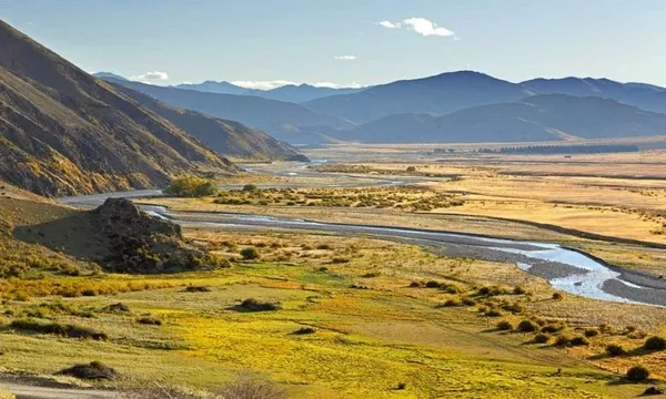 Sunny valley landscape with a winding river, grassy fields, and yellow patches. Hills rise on the left, mountains line the horizon, and the sky is mostly clear with a few clouds.