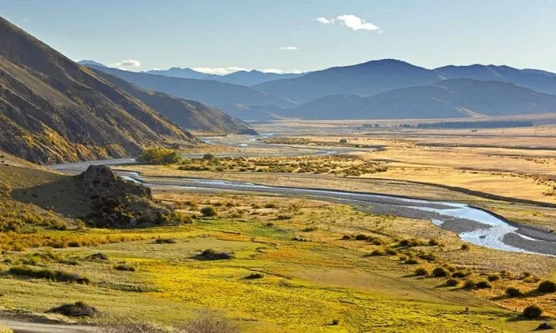 Sunny valley landscape with a winding river, grassy fields, and yellow patches. Hills rise on the left, mountains line the horizon, and the sky is mostly clear with a few clouds.