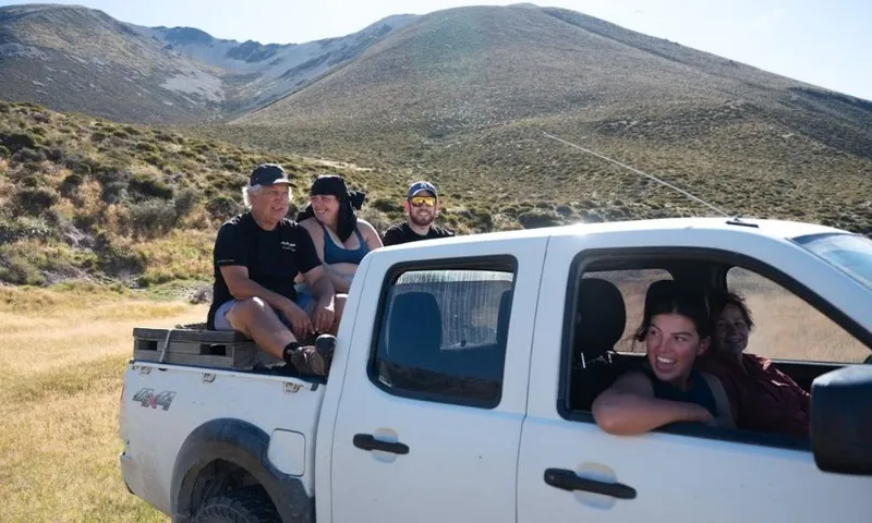 Five people enjoy an outdoor adventure; three sit in the bed of a white pickup truck, while two smile inside the cab, with grassy hills and a mountain in the background.