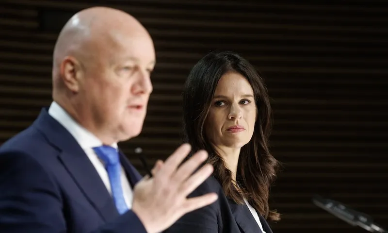 A man in a blue suit speaks while a woman with long brown hair, also in a dark suit, looks at him intently during a formal event or press conference.