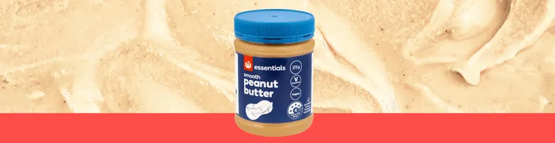 A jar of Essentials smooth peanut butter with a blue lid, placed in front of a background of creamy peanut butter and a red stripe at the bottom.