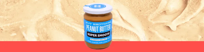 A jar of Nut Brothers Super Smooth Peanut Butter with a blue label sits on a red surface, against a creamy peanut butter background.