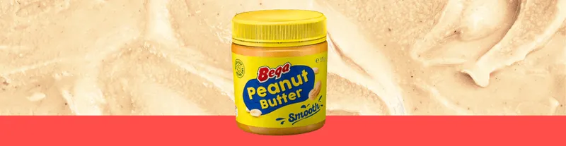 A jar of Bega Smooth Peanut Butter with a yellow lid and label is placed against a creamy peanut butter background with a red strip at the bottom.