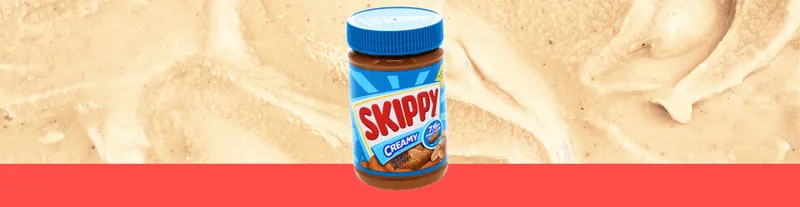 A jar of Skippy creamy peanut butter with a blue lid is centered on a background of smooth peanut butter and a red stripe at the bottom.