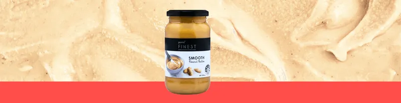 A jar of smooth peanut butter stands on a red surface with a creamy peanut butter texture in the background.