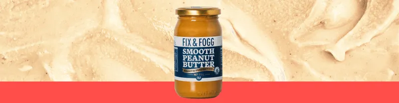 A jar of Fix & Fogg Smooth Peanut Butter stands against a background of creamy peanut butter spread with a pink border at the bottom.