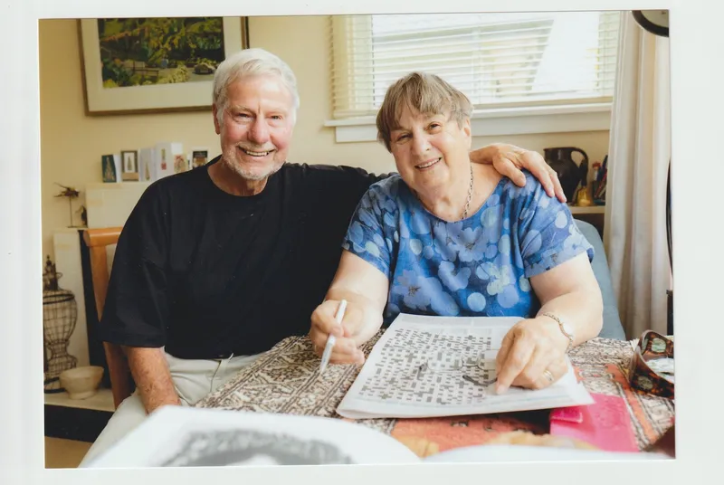 an older couple sit at a table doing a crossword and smiling at the camera. the man has his arm around the woman's shoulders