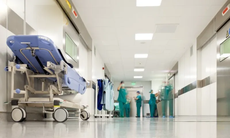 A hospital corridor with a stretcher to the left and medical staff wearing green medical scrubs in the background