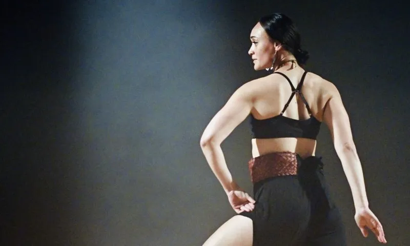 A dancer wearing a black outfit with a red belt performs on stage, facing sideways with one arm bent and leg slightly raised, under dramatic lighting.