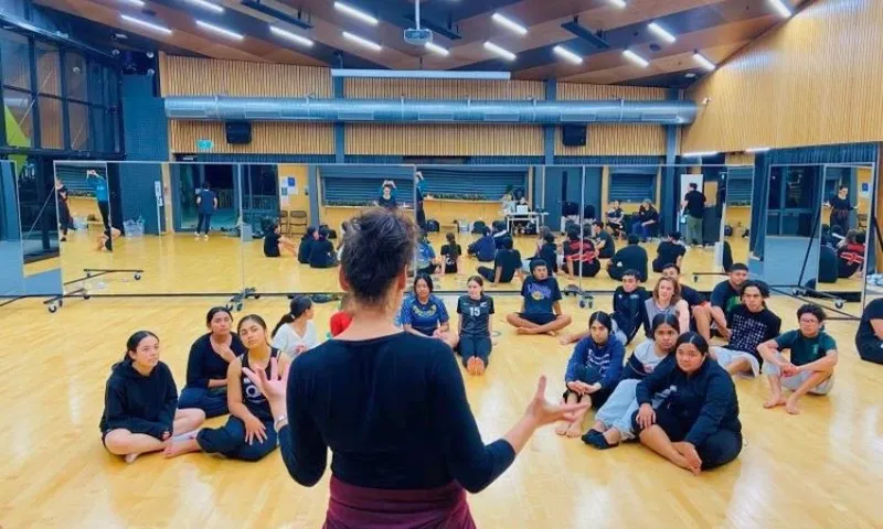 A person stands speaking to a group of people seated on a wooden floor in a mirrored dance studio. The group appears to be listening attentively. The room is spacious with modern lighting and large windows.