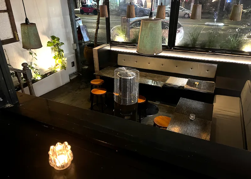 A dimly lit modern cafe with empty tables and chairs, illuminated by hanging lamps, a small candle on a table, and street view through large windows at night.