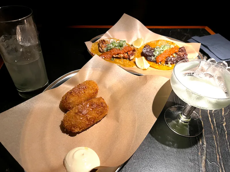 Two croquettes with dipping sauce, a cocktail, a drink with ice, and two tacos topped with red and green sauces are served on trays lined with brown paper on a dark table.