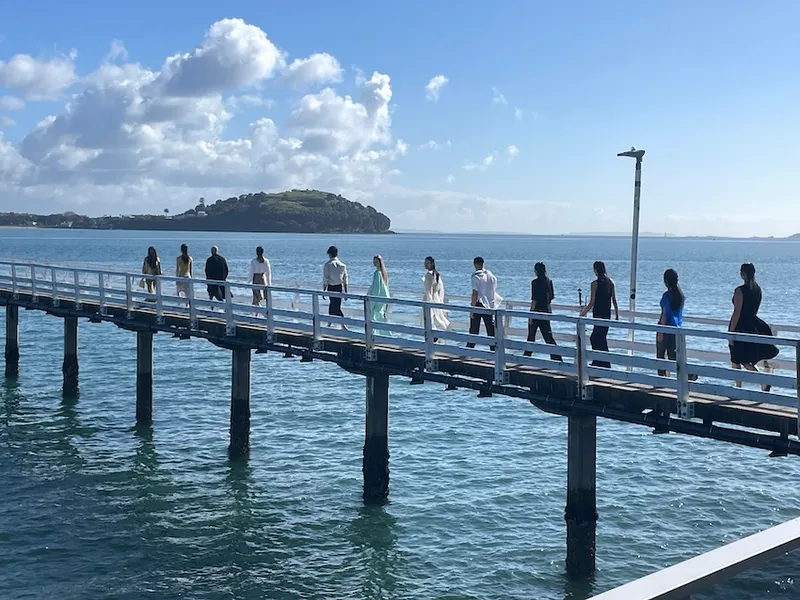 A group of people walk in a line across a long pier over calm blue water, with a green hill and partly cloudy sky in the background.