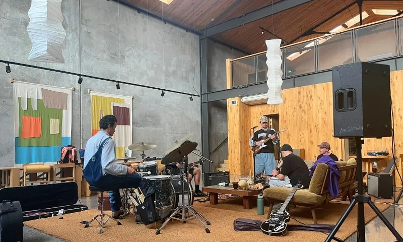 Four people practice music in a modern, loft-style room. One plays drums, another plays guitar, and two sit on a couch. A speaker, guitar, and colourful wall art are visible in the space.