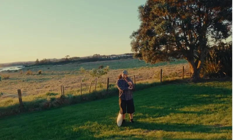 A person stands on a grassy lawn holding a shovel, with a large tree nearby and open fields stretching into the distance under a clear, golden sky.