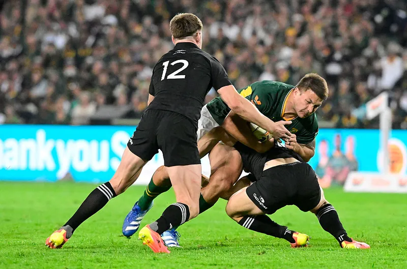 two men in black rugby uniforms tackle a man in a green uniform in front of a large crowd