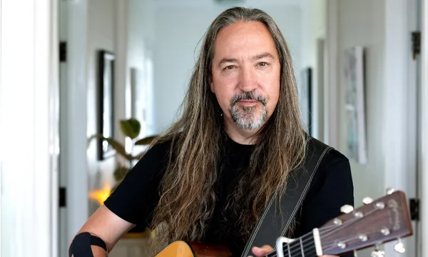 A photo of musician Jon Toogood who is standing in a hallway holding a guitar and looking at the camera. He has long hair and a beard.