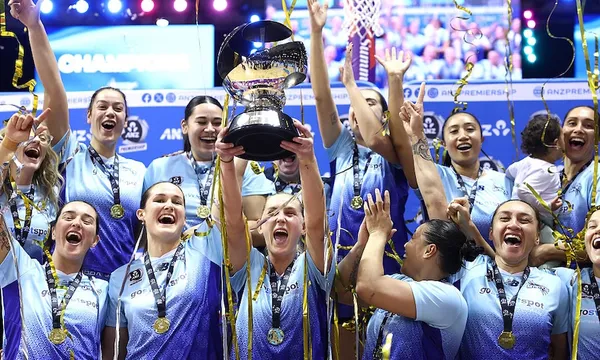 a close up group shot of a netball team of women in blue uniforms celebrating wearing medals and one holding a large trophy as streamers fall