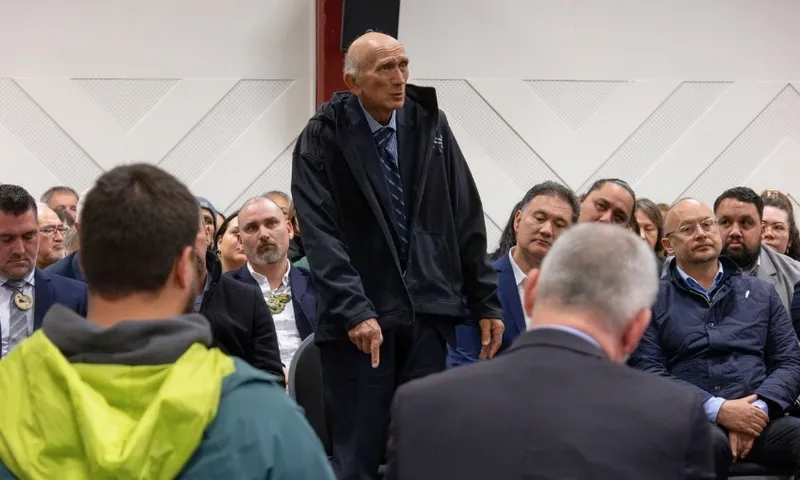 An older man in a black jacket stands and speaks among a seated group of people in formal and casual attire, gathered in what appears to be a meeting or conference room.