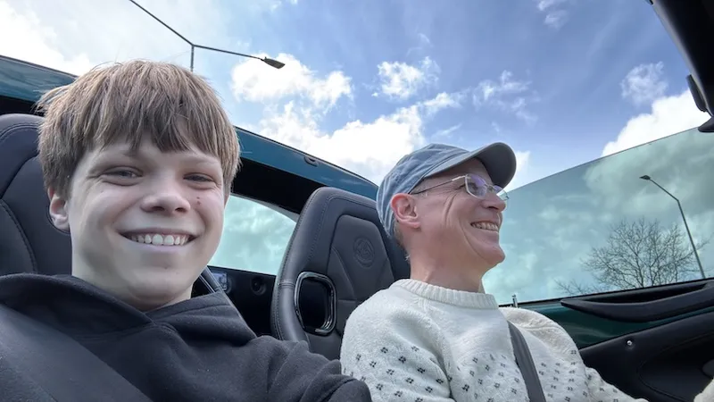 A smiling boy and a man in glasses and a cap sit in a convertible car with the roof down, enjoying a sunny day under a blue sky with scattered clouds.