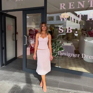 A woman in a light pink dress and sandals stands smiling in front of a boutique with glass doors and signage that reads “Designer Dresses.” There are pink and gold decorations visible inside the shop.