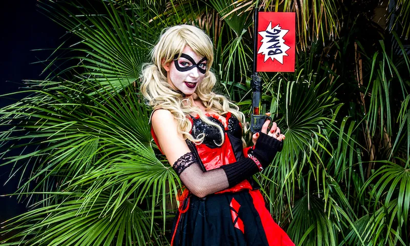 Portrait Of Woman In Harley Quinn Cosplay Costume Holding Gun With Bang Text Against Plants