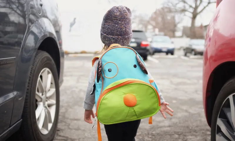 Rear view of young girl carrying bright green and blue backpack, walking in between parked cars.