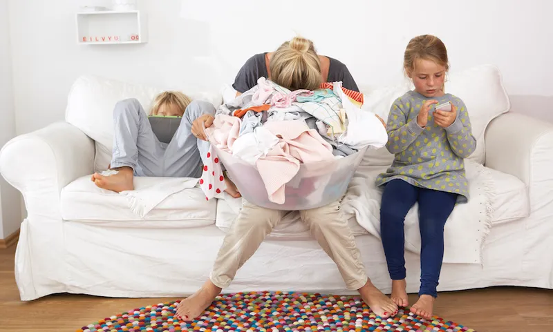 Exhausted mother with laundry basket on couch with children using digital tablet and cell phone