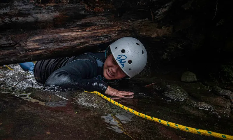 A student wearing a helmet crawls through a small gap in a rock, grinning and holding a rope.