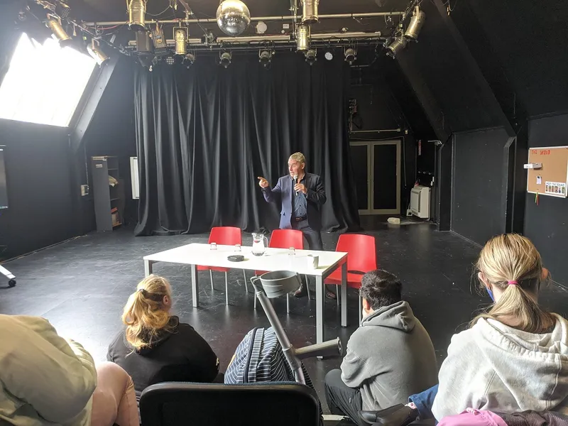 phil maguer in black room talking to people sitting on chairs who are looking at him