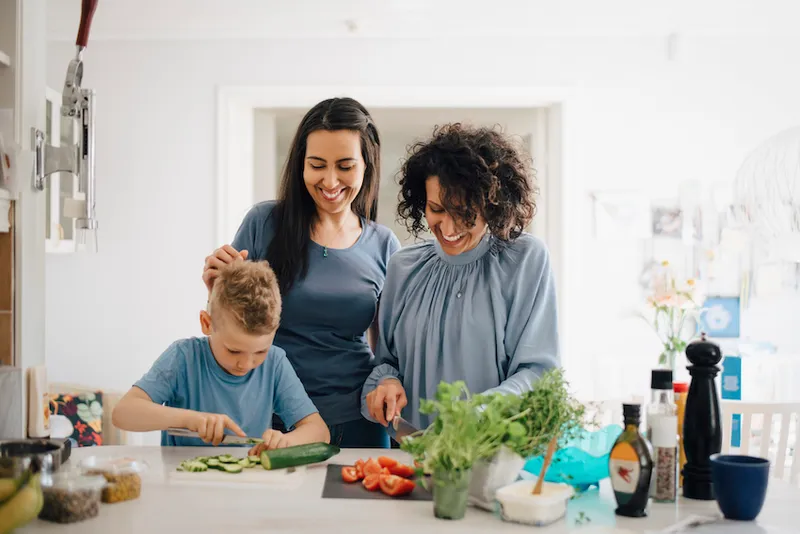 Smiling women looking at son cutting cucumber in kitchen