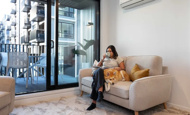 A woman is sitting on a sofa in a bright living room, sipping a drink and reading a book. Next to her is a dog resting comfortably on the couch. A sliding glass door opens to a balcony with outdoor furniture, showing apartment buildings outside.