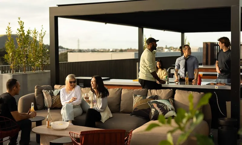A group of six people socializing on a rooftop terrace. Three are sitting on a couch with drinks, while the other three are standing and talking near an outdoor kitchen area. The background shows a scenic view of a cityscape.