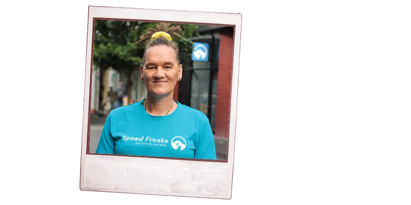Simone Winter with a yellow scrunchie in their hair and wearing a blue 'Speed Freaks' t-shirt is smiling and standing on a city street.