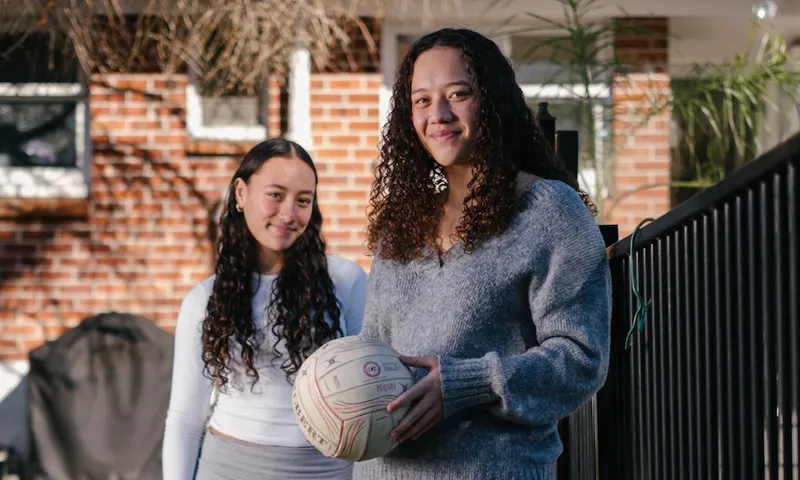 Two girls stand outside their red brick home, they have a volleyball wth them, both are smiling, have brown curly hair worn down and are wearing white tops. The are both smiling.