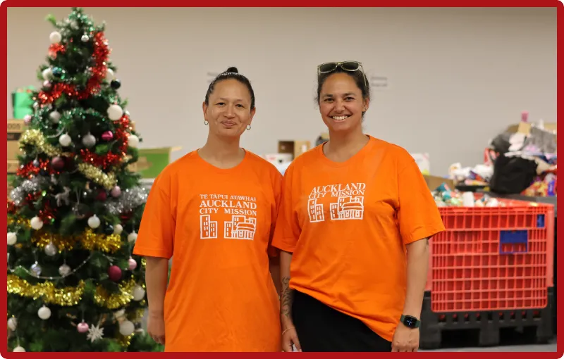 Anataia Fu and Tara Moala stand side by side in orange Auckland City Mission t-shirts. They are standing in front of a Christmas tree with tinsel and many white and red baubles. They are both smiling and there are boxes behind them which are waiting to be packed full of gifts to share with families.