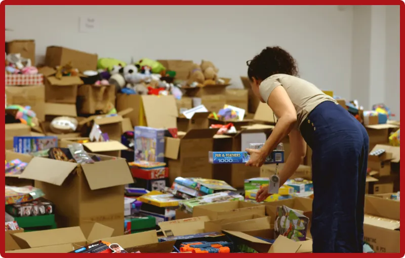 A volunteer in a beige top, dark blue jeans, selects a few toys from an area full of carboard boxes full of books, toys, games and other holiday gifts.
