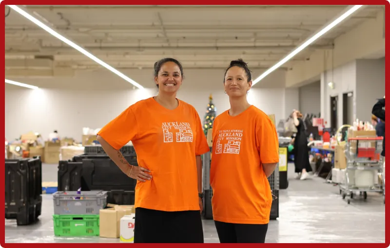 Two women, Tara Moala at left and Anataia Fu at right, wear orange Auckland City Mission tshirts and black pants and are standing side by side smiling in front of the gift and food packing stations in a large open warehouse space.