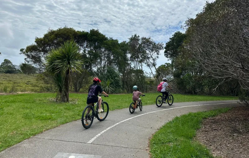 Three people of various ages ride bicycles on a paved path through a park. They are wearing helmets and surrounded by green trees and grass under a cloudy sky.