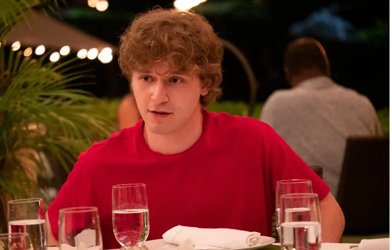 A person with curly hair wearing a red shirt sits at a table surrounded by several glasses of water, appearing to be engaged in conversation. The background is softly lit and includes plants and a partially blurred person.