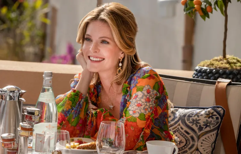 A woman in a colorful floral dress smiles while sitting at an outdoor table set with glassware, a bottle of water, and food. There is a plant in the background.